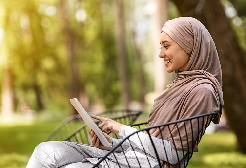 Attractive arab woman in headscarf using digital tablet, sitting on bench at park, side view, copy space