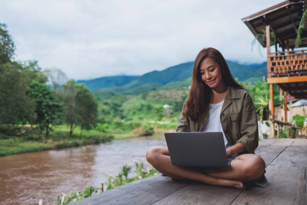 a beautiful asian woman working on laptop computer while sitting by the river with mountains and nature background