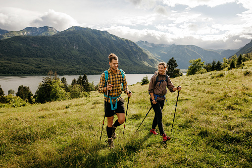 Young adult couple hiking on a hill over Lake Bohinj in Slovenia. They are wearing hiking clothes, backpacks and holding hiking poles. Horizontal photo with copy space. Full lenght body. Bright sunny day with lake in the background.