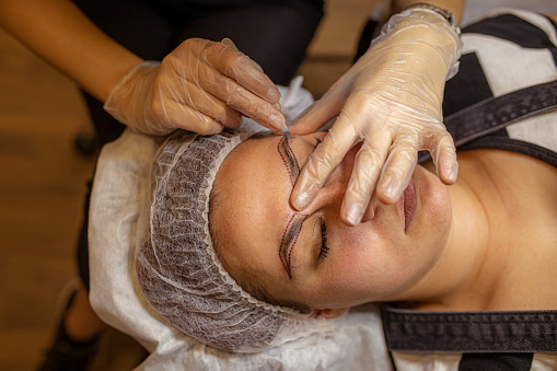 Closeup of beautician preparing young woman for a permanent makeup treatment of eyebrows
