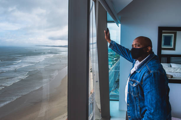 Man with a face mask looking out to empty beach and quiet roads during lockdown An African man with a black face mask and denim jacket being frustrated at home during lockdown. He is looking out through a window down to empty beach and quiet roads during lockdown. black face mask side view stock pictures, royalty-free photos & images