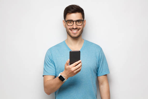 istock Young handsome man in blue t-shirt holding smartphone and looking at camera with smile while standing isolated on gray background 1257722803