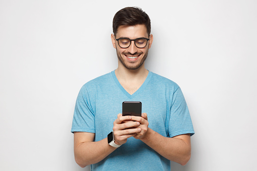 Portrait of young handsome guy wearing blue-shirt and glasses looking at phone screen with smile, isolated on gray background