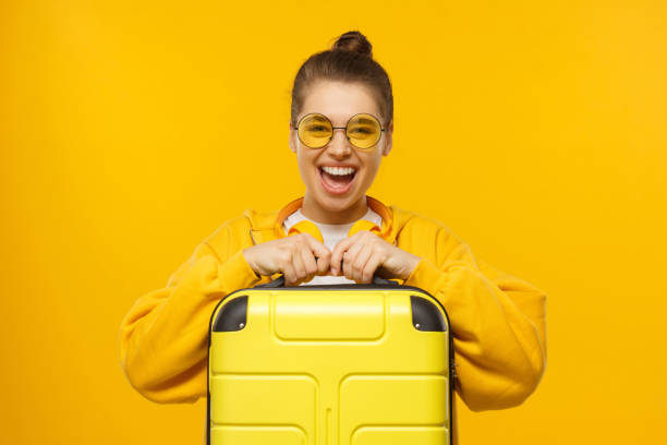 fille heureuse excitée d’adolescence utilisant le hoodie, les glaces colorées et les écouteurs autour du cou, retenant la valise de voyage, prête pour le vol, isolée sur le fond jaune - luggage hold photos et images de collection