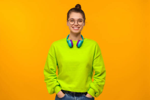 녹색 네온 스웨트셔츠와 청바지를 입은 어린 행복한 소녀, 목에 무선 헤드폰을 쓰고, 주머니에 손을 대고, 노란 배경에 고립되어 있습니다. - female likeness audio 뉴스 사진 이미지