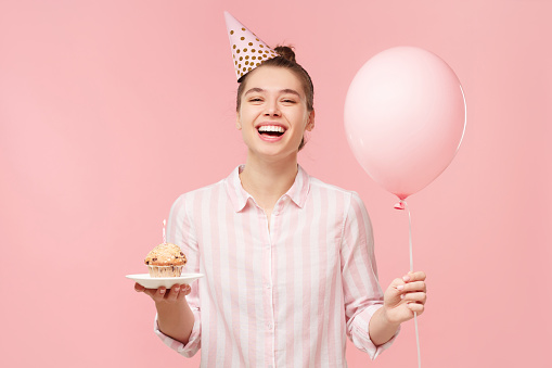 Beautiful smiling woman in dress and birthday hat holding gift box and pastel pink air balloons isolated on pink background. cute happy young girl on a birthday party. copy space