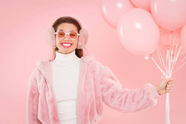 young happy excited girl wearing ear warmers and colored glasses, holding birthday balloons, isolated on pink background - personal accessory balloon beauty birthday imagens e fotografias de stock