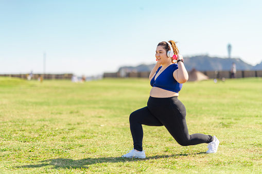 A young female athlete is doing muscle trainings with dumbbells at a public park on grass.