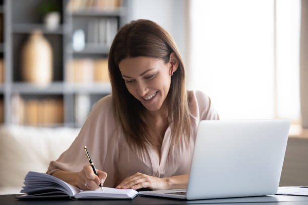 mujer sonriente escribiendo en el cuaderno, usando el ordenador portátil en casa - lecture notes fotografías e imágenes de stock