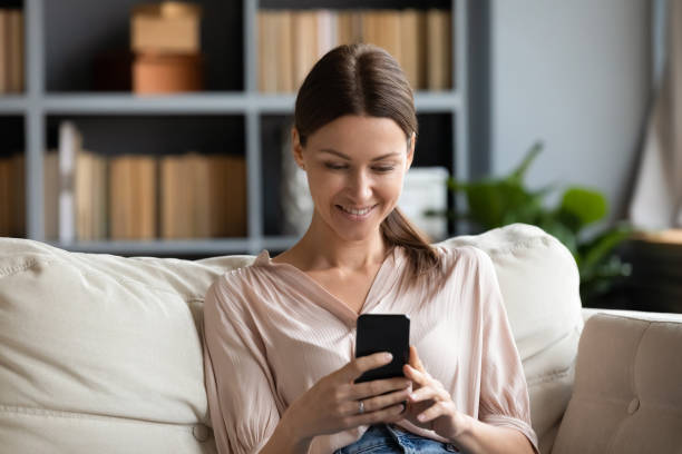 istock Smiling young woman using phone, sitting on couch at home 1257750304