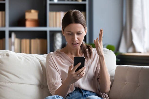 Confused angry young woman having problem with phone Confused angry woman having problem with phone, sitting on couch at home, unhappy young female looking at screen, dissatisfied by discharged or broken smartphone, reading bad news in message poor signal stock pictures, royalty-free photos & images