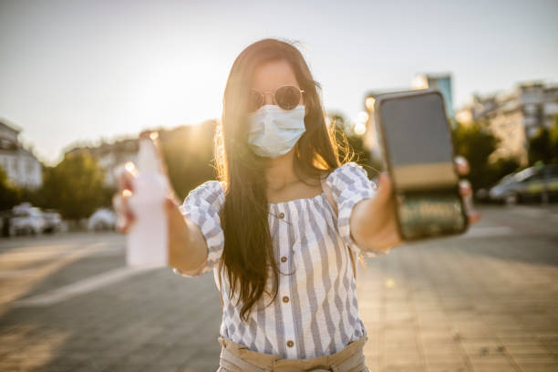istock Woman wearing a surgical mask, disinfecting her mobile phone before using and standing on empty plaza during the lockdown 1257752211