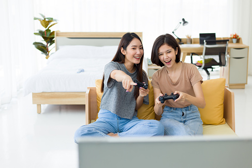 Two young Asian women holding game joysticks and competing happily while sitting on the sofa. Competitive friends playing video games at home.