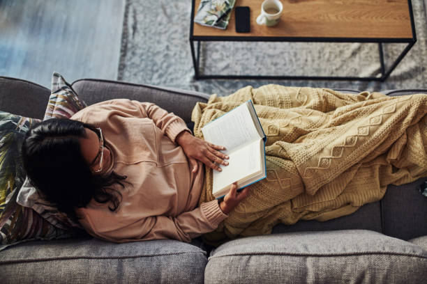 A good book can do the world of good High angle shot of a young woman reading a book on the sofa at home cozy stock pictures, royalty-free photos & images