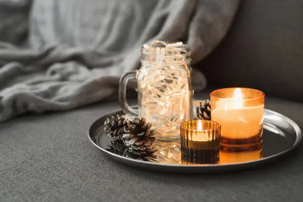 Photo of Living room décor: two burning aroma candles of orange color, pine cones and Christmas lights in a glass jar. Hygge concept. Autumn - Winter season