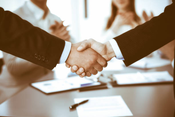 istock Business people shaking hands at meeting while theirs colleagues clapping and applauding. Group of unknown businessmen and women in modern white office. Success teamwork, partnership and handshake concept 1257761973