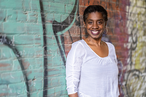 Portrait of a beautiful African American woman in her 50s leaning up against a graffitied wall looking at the camera.