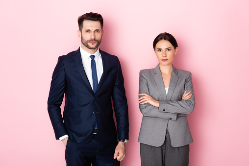 handsome businessman standing with hand in pocket near businesswoman with crossed arms on pink, gender equality concept