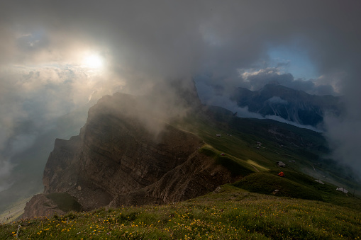 Mount Seceda view, at sunrise with clouds and fog and colorful bloomed flowers in the foreground, Dolomites Italy