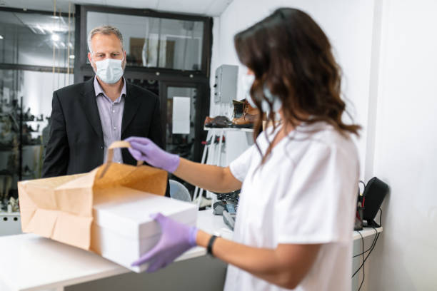 cliente che indossa una maschera protettiva per il viso in attesa che l'addetto alle vendite imballi la scatola delle scarpe - shoe store sales clerk customer foto e immagini stock