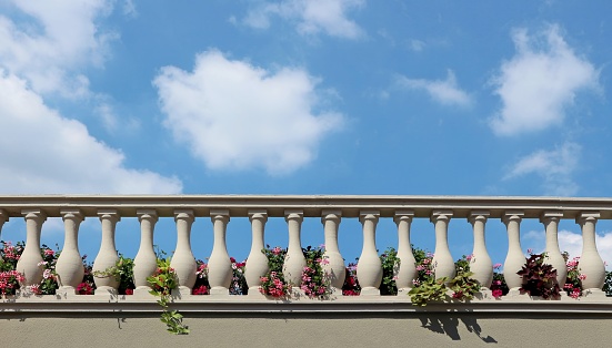 Stone balustrade of a balcony with flowers between the columns. Blue sky with fluffy clouds on background. Copy space