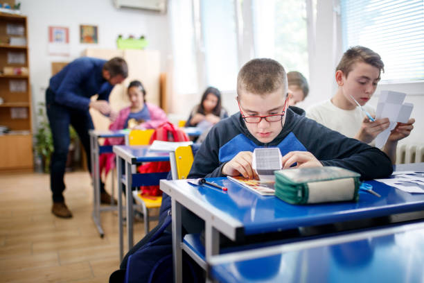 Focused special needs boy folding paper Intellectual disabiled elementary age boy is folding paper following pattern, his deskmate is cutting paper using scissors, their teacher is in background special education stock pictures, royalty-free photos & images