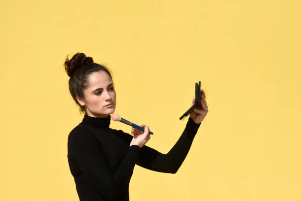 istock Side view of a young woman with ponytail using the phone as a mirror while applying makeup brush on face skin 1257788080
