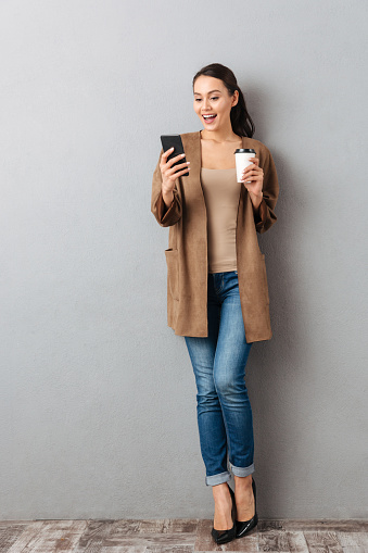 Full length of a cheerful young asian woman using mobile phone while standing and holding cup of coffee over gray background