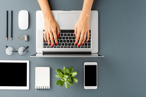 Overhead shot of woman’s hands working on laptop on gray table with accessories. Office desktop. Flat lay
