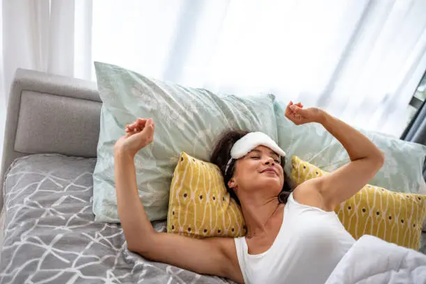 Morning woman waking up stretching in bed in sunshine sunrise early bird happy girl in pajamas on weekend in sun glow. Healthy wake-up routine home lifestyle.