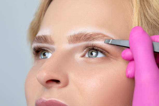 istock Woman with blue eyes having Permanent Make-up Tattoo on her Eyebrows. Eyelash artist plucks eyebrows with tweezers. Professional makeup and cosmetology skin care. 1257809756
