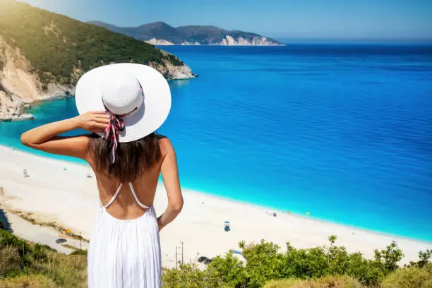 A beautiful tourist woman in white dress gazes the famous beach of Myrtos, Kefalonia, Greece, with turquoise blue sea and sunshine during summer time