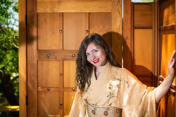 Young woman happy girl smiling face in kimono standing by wooden door in modern outdoor garden zen home Japanese Japan architecture