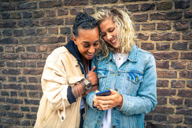 Happy girlfriends looking at a smartphone and having fun - Multiracial lesbian couple, millennials women, having fun together in London - LGBTQ concept with mixed race beautiful couple