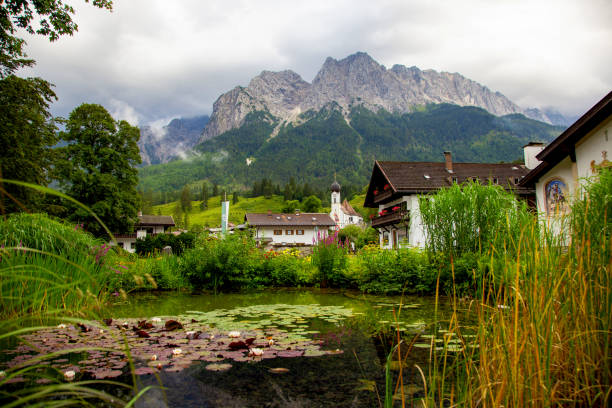 grainau, villaggio bavarese. chiesa a cupola st. johannes, cimitero con le montagne (waxenstein e cime di zugspitze). wetterstein gamma northern limestone alpi bayern germania europa. stagno in primo piano - grainau foto e immagini stock