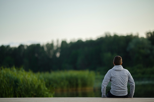 Young man sitting alone on edge of footbridge at lake in summer calm evening. Thinking about life. Spending time alone in nature. Peaceful atmosphere. Back view.