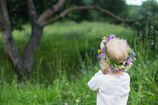 Small girl with blond curly hair wearing colorful wreath made of various wild flowers looking at pine tree. Backview. Happy summer mood