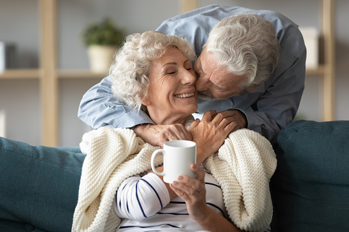 Loving older husband kissing smiling wife on cheek, covering warm plaid, expressing love and care, aged senior couple enjoying tender moment, mature woman holding cup of tea coffee, relaxing on sofa