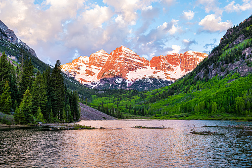 Maroon Bells in Aspen, Colorado with clouds in blue sky at sunrise and rocky mountain peak lake reflection in summer with red sunlight and green forest trees
