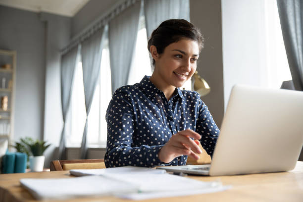 istock Smiling indian girl sitting at table, looking at laptop screen. 1257836726