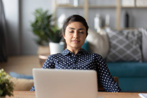 Happy peaceful millennial indian girl leaning on chair, resting. Happy peaceful millennial indian girl leaning on chair, napping, enjoying free break pause time at home. Calm young hindu woman relaxing with closed eyes, sitting alone at desk with computer. mindfulness photos stock pictures, royalty-free photos & images