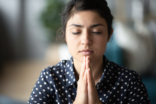 Head shot close up millennial hindu girl student folding hands, praying god for good luck. Worried religious young indian woman asking for help, waiting for miracle or feeling thankful alone at home.