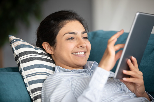 Head shot close up happy millennial indian woman lying on sofa, chatting online with friends e-dating in social networks using digital tablet. Cheerful young girl enjoying web surfing shopping online.