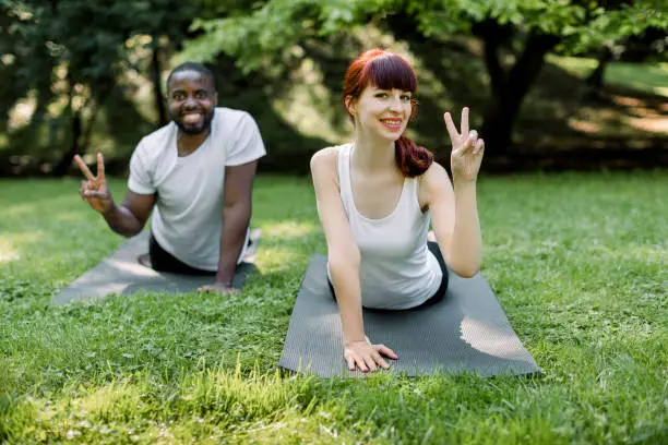 Happy multiethnic fitness couple practicing yoga performing yoga-asanas, laying and stretching on a yoga mat outdoors, smiling at camera and showing victory sign. Focus on woman.