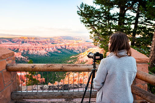 Woman taking pictures photos of view from Bryce Point overlook of hoodoos rock formations in Bryce Canyon National Park at sunset with tripod and camera