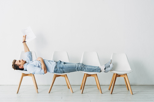 Side view of employee holding resume while lying on chairs in office