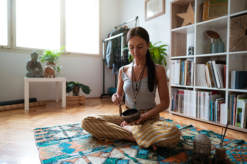 Young Caucasian woman sitting in a lotus position with eyes closed, using meditation bell to relax while meditating and doing breathing exercises at 
home.
