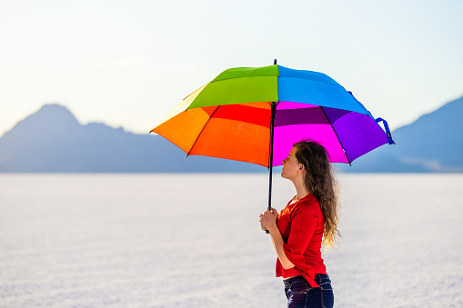 Young woman holding colorful rainbow umbrella for shade on white Bonneville Salt Flats near Salt Lake City, Utah and mountain view