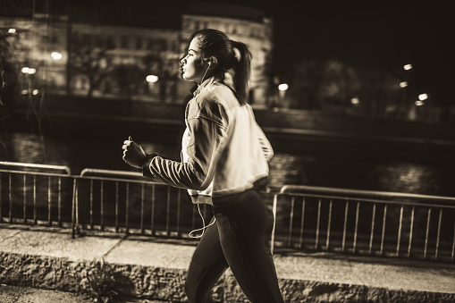 Young sportswoman with headphones running on sidewalk at night