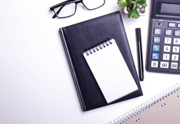 istock On a white table are a calculator, a black pen, a notebook and a flower in a pot. Top view close up, place for text. 1257893646